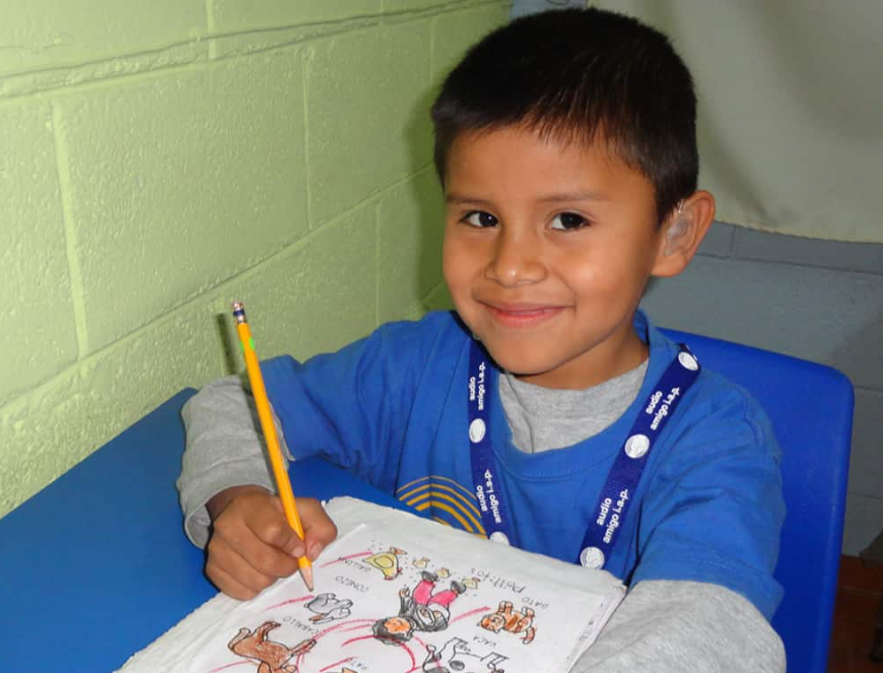 Niño con aparato auditivo dibujando con un lápiz en clase
